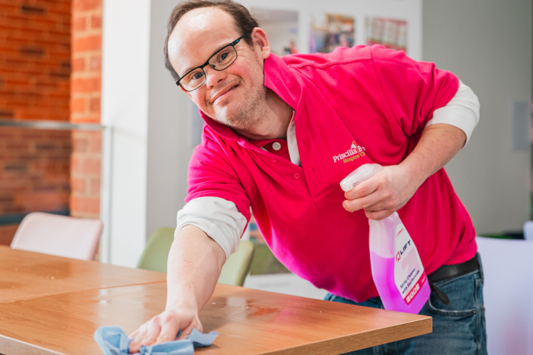 The image shows a young man with down syndrome wearing a bright pink polo shirt. He is holding a cleanign spray bottle in one hand, and some blue roll in the other and he is using the blue roll to wipe a table which is in front of him.