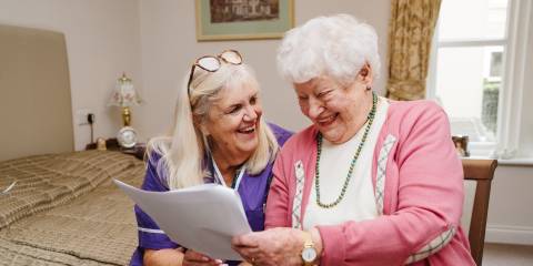 A care worker and elderly lady are sitting together looking at a piece of paper and smiling, the elderly lady is looking at the piece of paper and the care worker is smiling at the elderly lady.