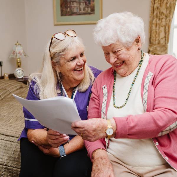 A care worker and elderly lady are sitting together looking at a piece of paper and smiling, the elderly lady is looking at the piece of paper and the care worker is smiling at the elderly lady.
