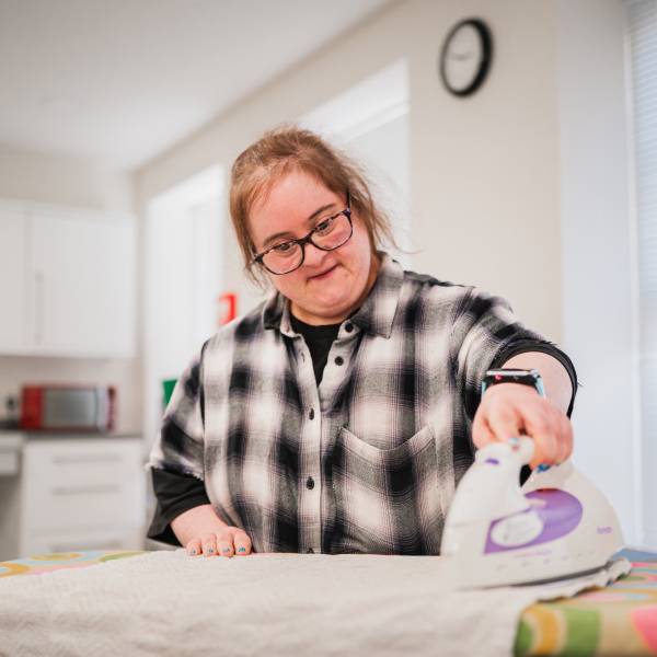 The image shows the torso, head and shoulders of a lady with learning disabilities stood in front of an ironing board, holding the iron in her left hand and ironing a towel.