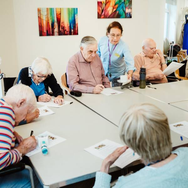 The photo shows a large table with five elderly people sat around it with pieces of paper in front of them, some looking at the paper and some writing. There's one care worker standing behind them and there is colourful art on the wall behind them all.