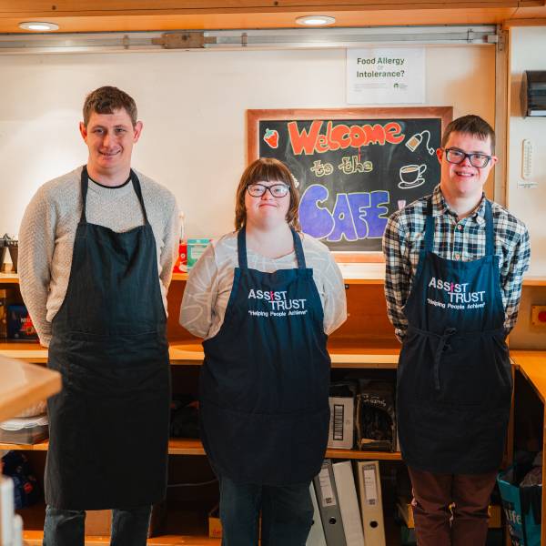 The image shows individuals with learning disabilities standing, all wearing aprons, with a sign behind them that says "Welcome to the Cafe"